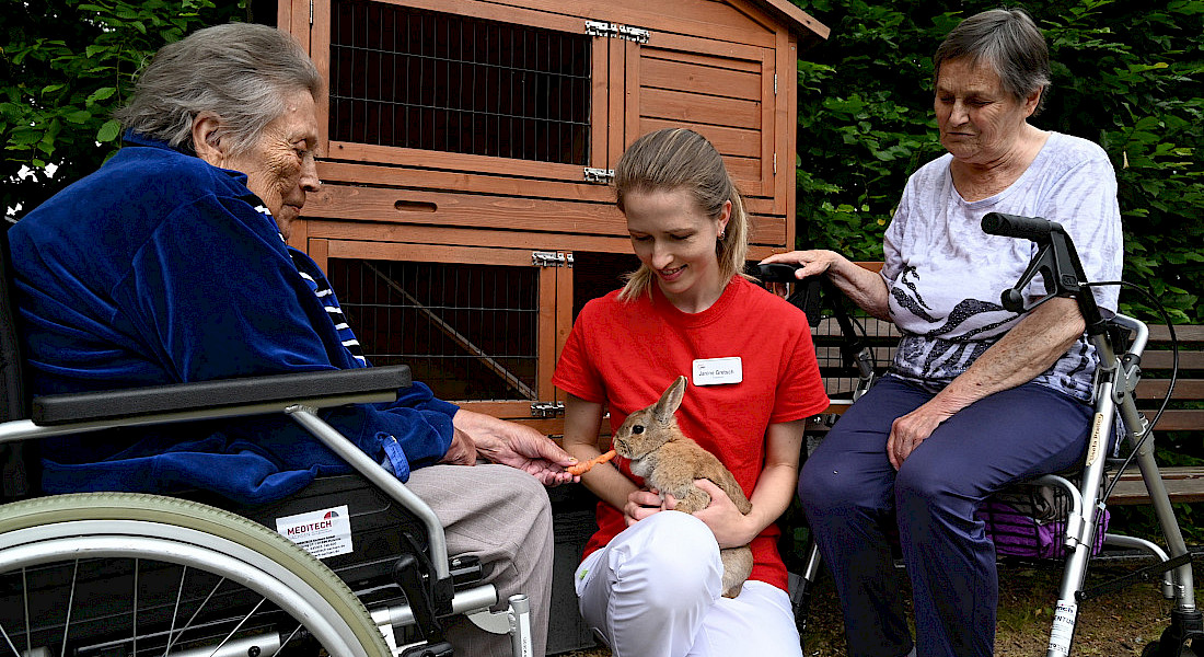 Eine Pflegekraft kniet neben zwei Personen im Rollstuhl und mit Rollator, die Pflegekraft hält ein Kaninchen welches gestreichelt wird.