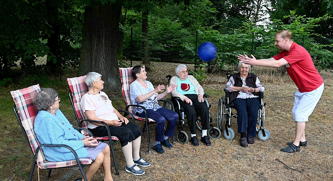 Gruppe älterer Menschen sitzt im Schatten unter Bäumen, eine Pflegekraft wirft ihnen einen Ball zu.