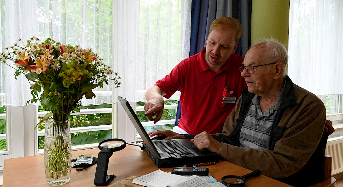 Zwei Personen sitzen an einem Tisch, eine Pflegekraft unterstützt die bei der Nutzung eines Laptops.