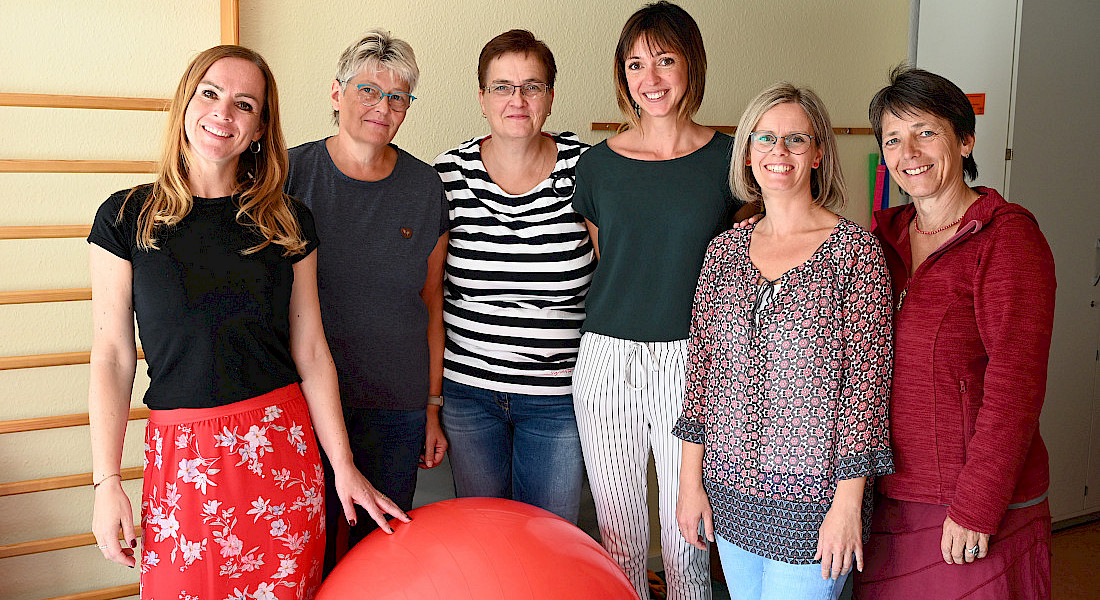 Sechs Personen stehen nebeneinander und lächeln in einem Raum mit Gymnastikmaterialien.