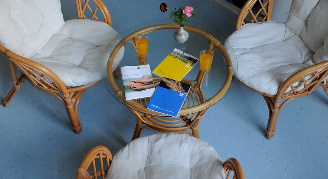 Drei Stühle um einen Glastisch. Auf dem Tisch liegen verschiedene Infomaterialien und Flyer aus.
