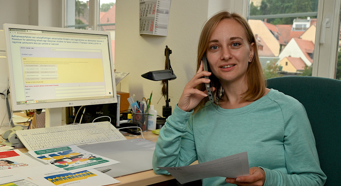 Einen Frau telefoniert und hält einen Zettel in der Hand. Sie sitzt am Schreibtisch und schaut in die Kamera.