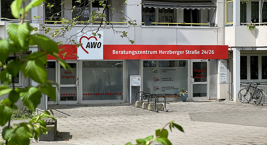 Blick an einem Ast vorbei auf das AWO Beratungszentrum, ein rotes langes Banner ist über dem Eingangsbereich, mehrere Fahrradständer sind vor dem Eingang.