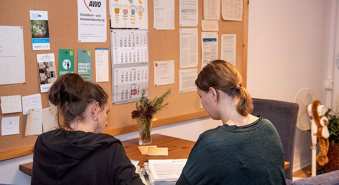 Zwei Personen sitzen an einem Tisch vor einer Pinnwand mit Aushängen und sprechen miteinander.