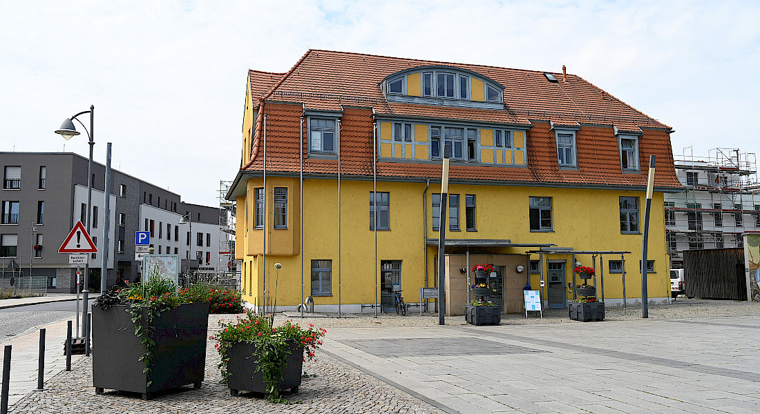 Freistehendes, gelbes Gebäude mit rotem Dach an einem gepflasterten Platz.
